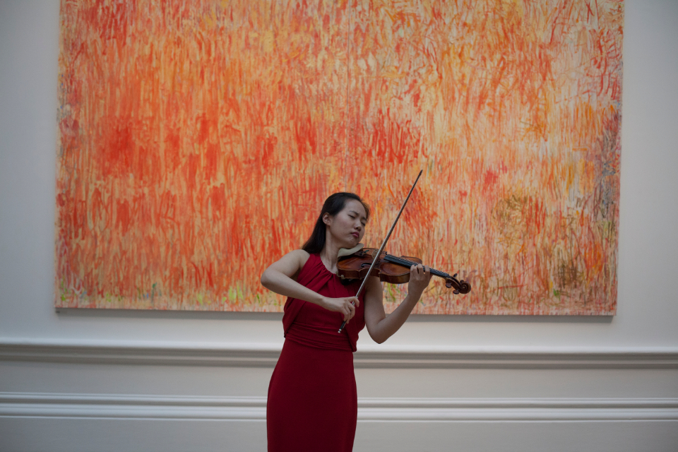 An Asian student, wearing a red dress, performing on the violin, with an art piece behind here.